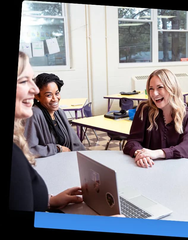 Three women sitting around a table in a classroom, smiling and engaged in conversation, with a laptop open on the table.