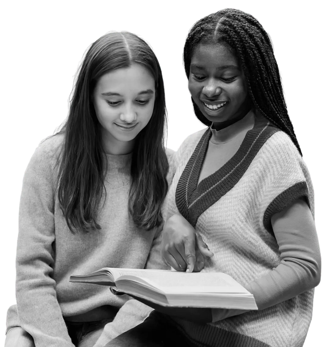 Two young women sitting together and reading a book, one pointing at the text and both smiling.