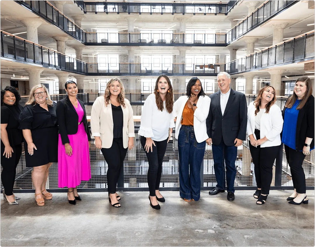 Group of eight women and one man standing indoors in a modern multi-story building, all smiling and dressed in business casual attire.
